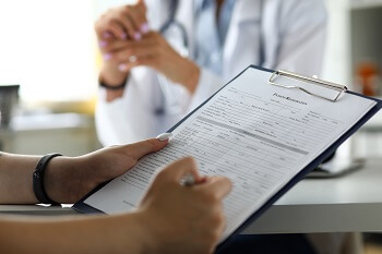 Patient filling out medical forms on a clipboard during a consultation with a doctor in a clinical setting